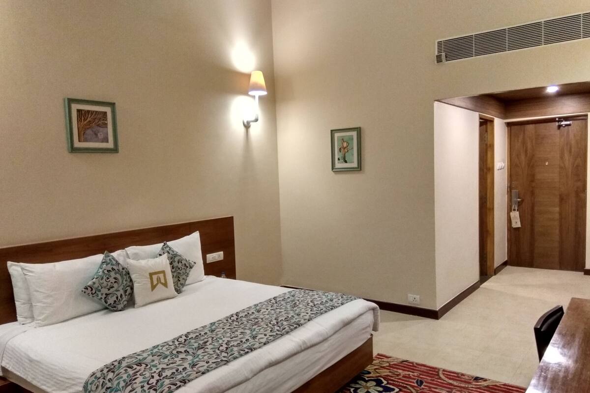 Habitación de hotel con cama, escritorio, silla, lámpara y dos cuadros en la pared.