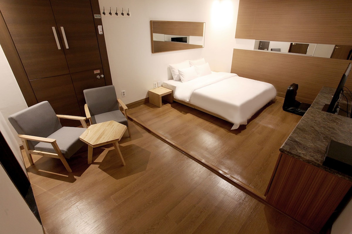 A hotel room with a bed, two chairs, a small table, a nightstand, a mirror, and a wooden wardrobe.