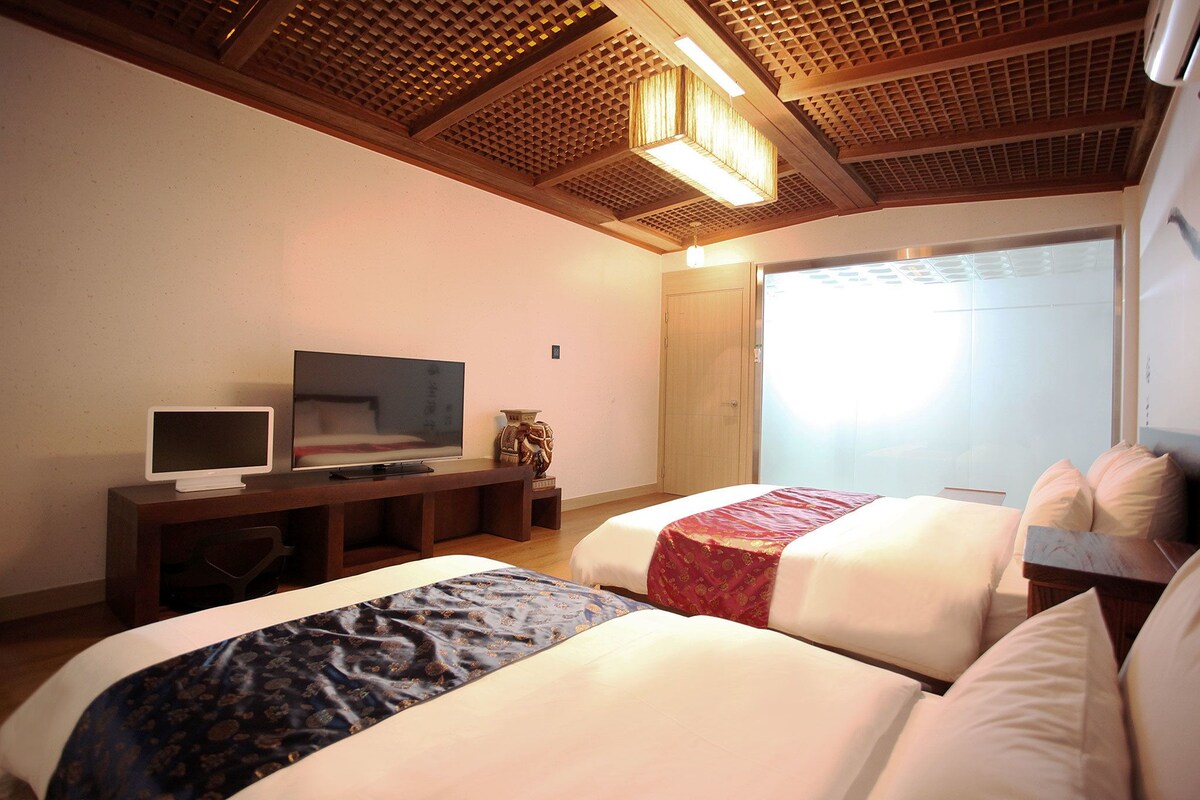 A hotel room with two beds, a TV, a desk, and a window with blinds.