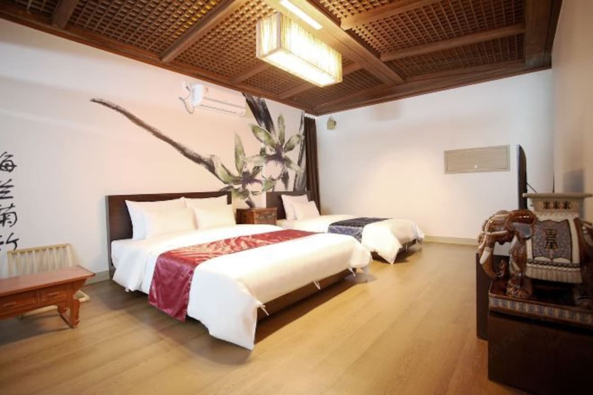 A hotel room with a large bed, two bedside tables, a wooden floor, a ceiling fan, and a wall-mounted air conditioning unit.