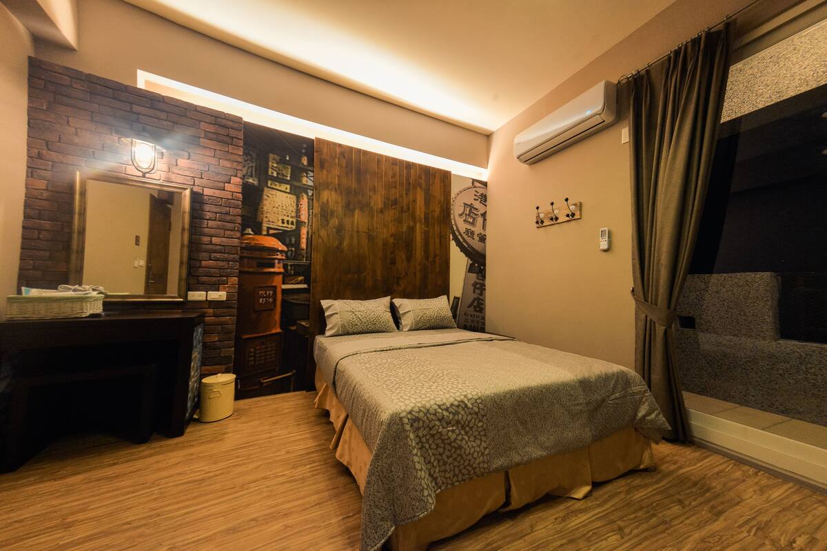 A hotel room with a bed, a desk, a mirror, a wooden wall, and a window with curtains.