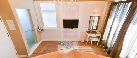 A bedroom with a wooden floor, a vanity with a mirror, a television on the wall, and a window with blinds.