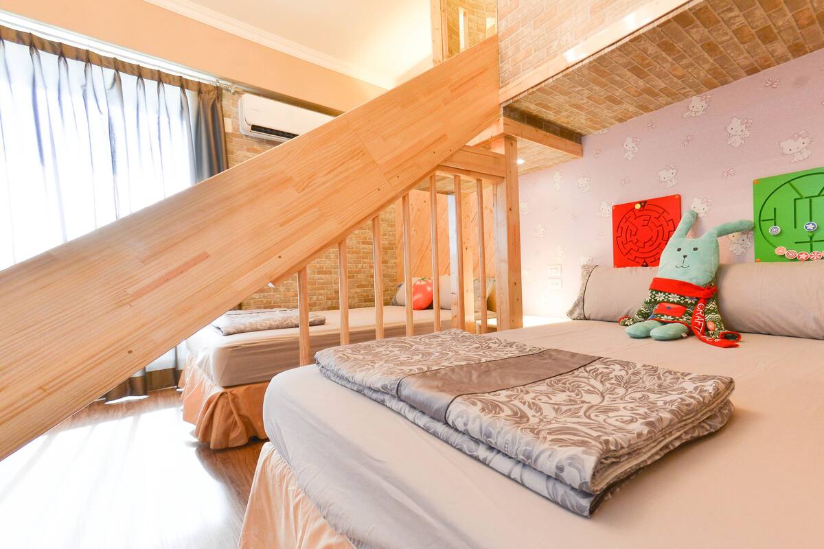 A bedroom with a sloped ceiling, a wooden staircase, a bed with patterned bedding, a plush toy, and a wall with Hello Kitty decorations.