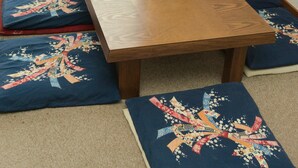 Une table à manger en bois, ornée d’une horloge, de coussins à motifs floraux bleus, et offrant une vue sur un bâtiment au toit bleu, visible à travers une fenêtre.