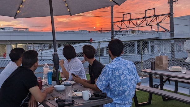 Un groupe de personnes est assis à une table en plein air, à l'ombre d'un grand parasol, et profite d'une vue sur la ville au coucher du soleil.