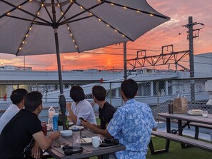 A group of people dining at an outdoor table with a large umbrella, overlooking a cityscape with a sunset. - Guest House Kyoto Inn - Hostel (Kyoto)