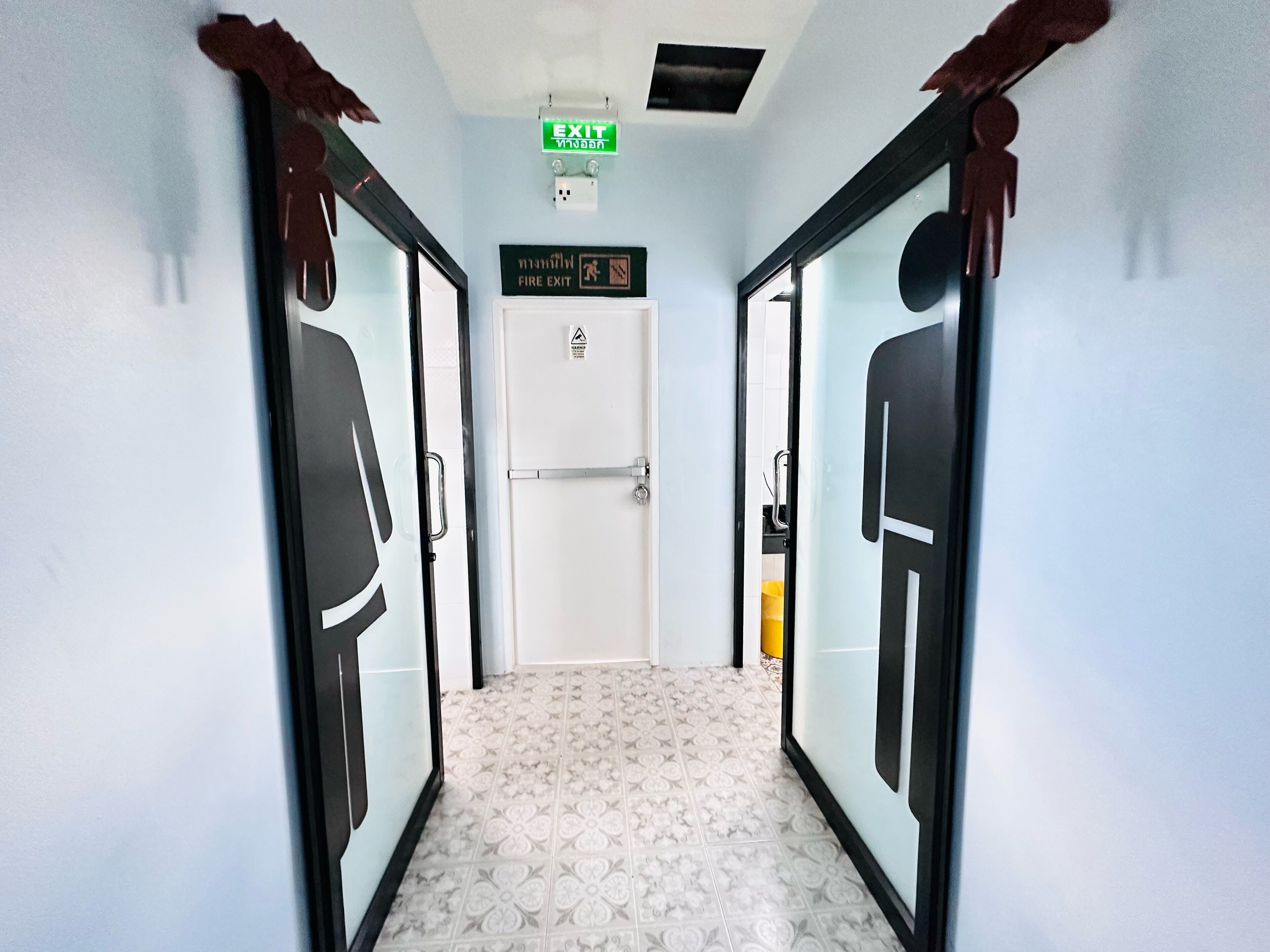 A hallway with patterned floor tiles, a white door, and two doors with black frames and glass panels. An EXIT sign is visible above the door.