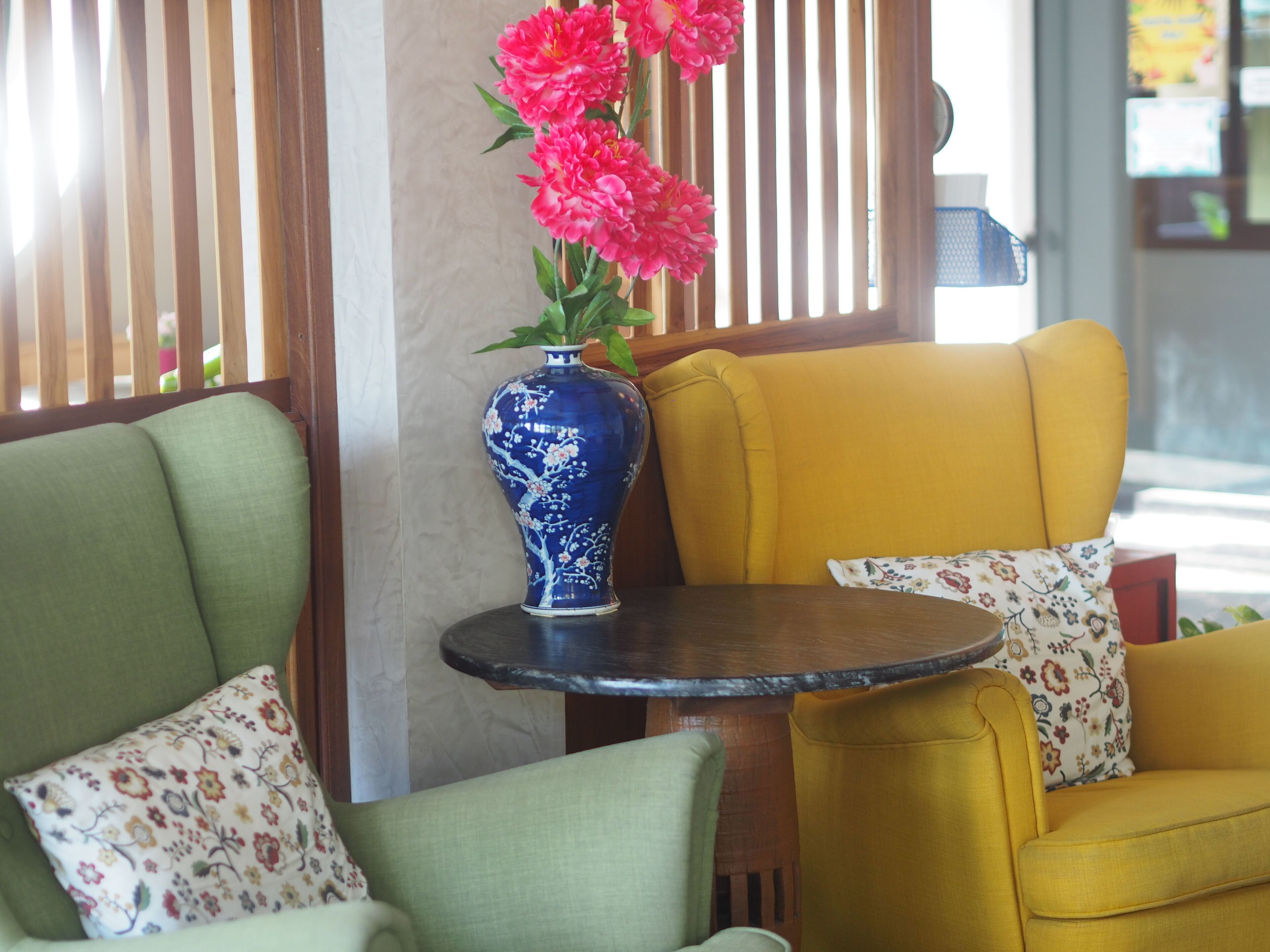 Quarto com um vaso azul com flores cor-de-rosa sobre uma mesa redonda pequena, cadeiras verdes e amarelas, e almofadas com padrão floral.