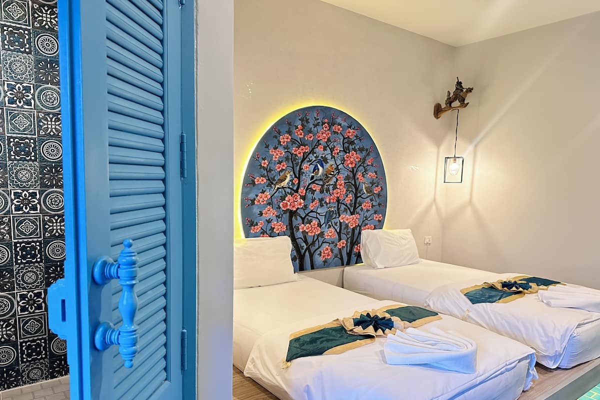 Habitación de hotel con dos camas, un mural decorativo, una puerta azul y un salpicadero de azulejos estampados.