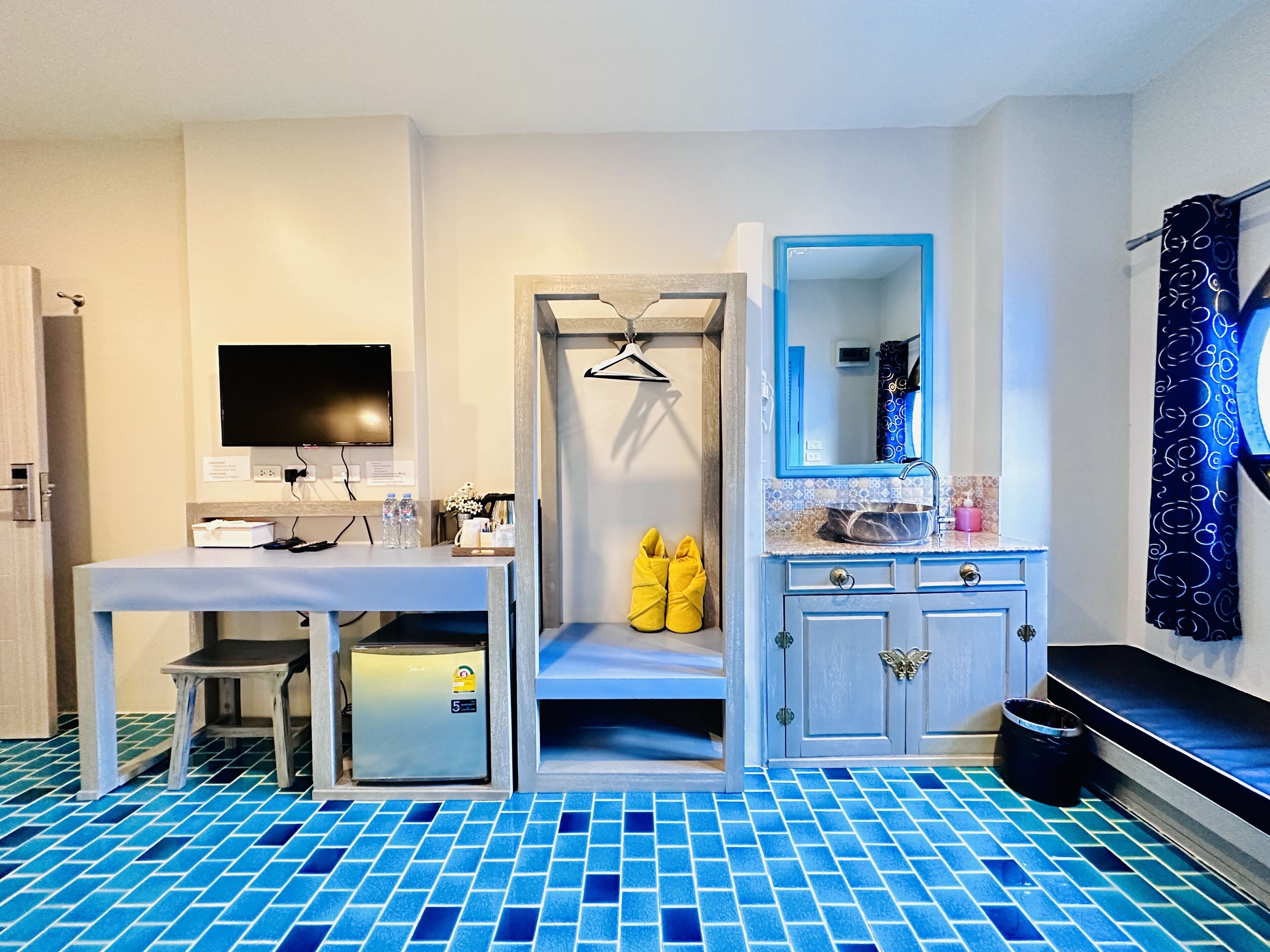 A hotel room with a tiled floor, a desk with a computer, a mirror, and a small seating area with yellow slippers.