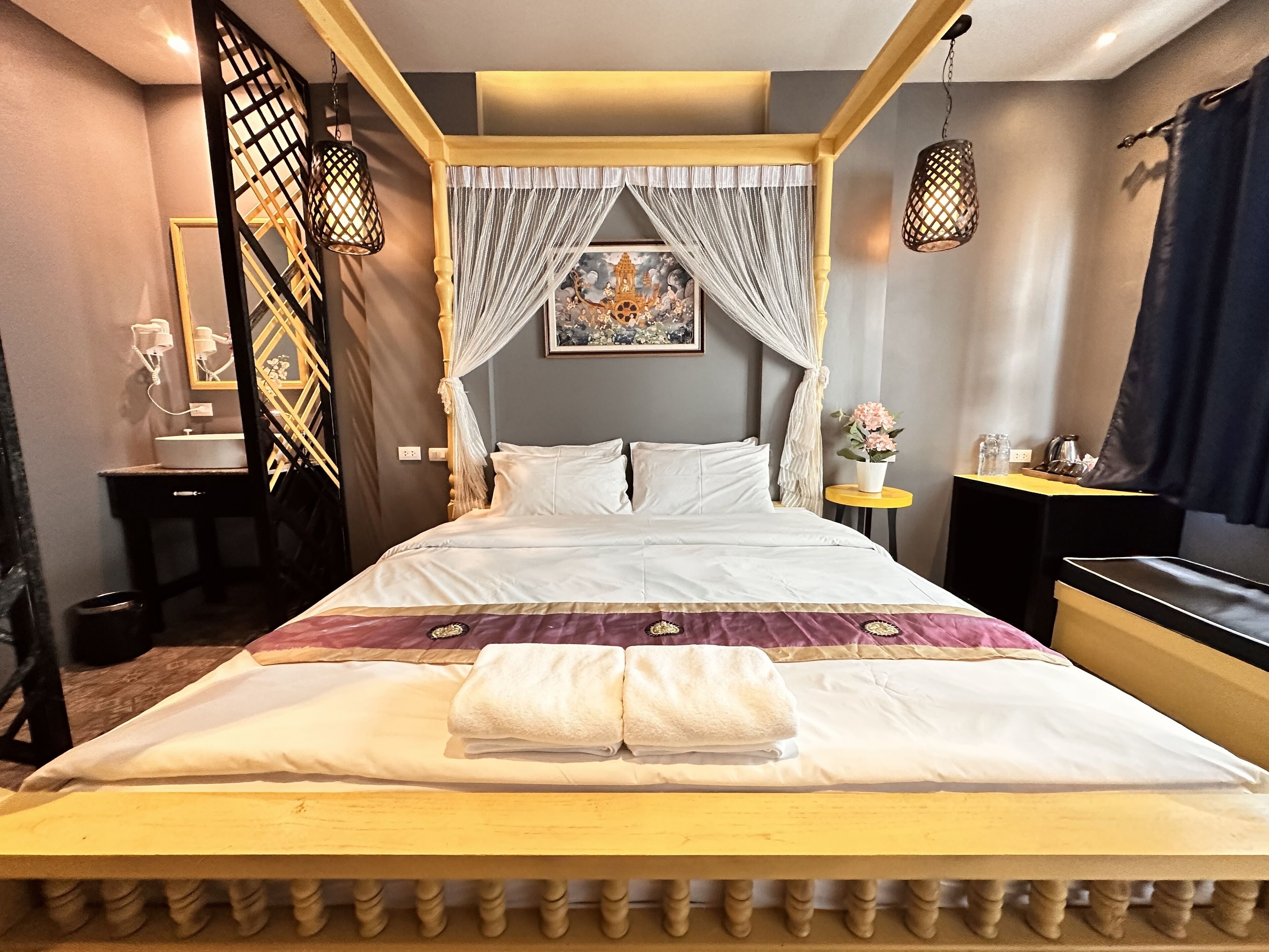 A neatly made bed with a canopy, a bedside table with a lamp, and a small table with a flower vase.