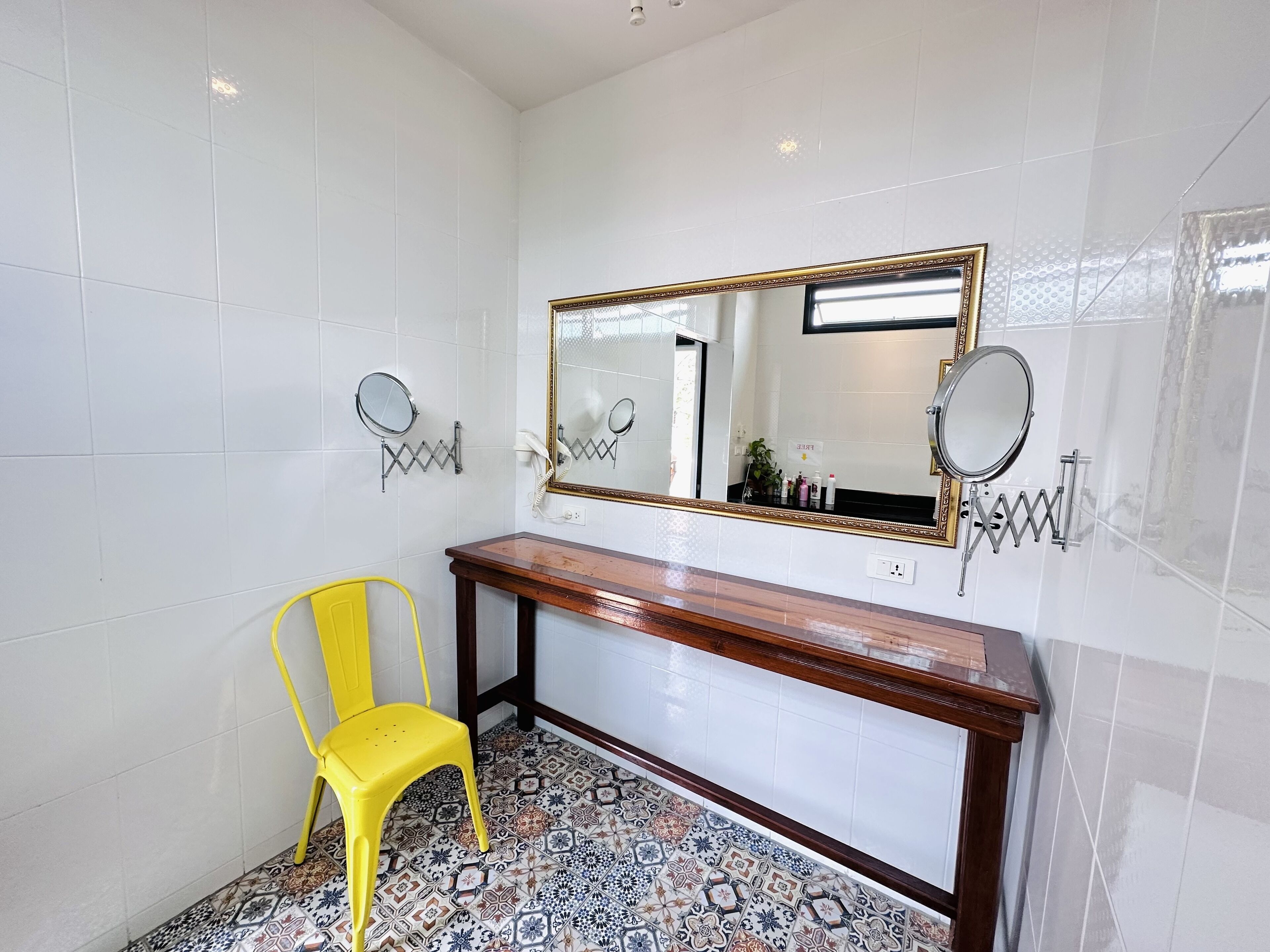 A bathroom with white tiled walls, a wooden vanity, a yellow chair, and a large mirror with a gold frame.