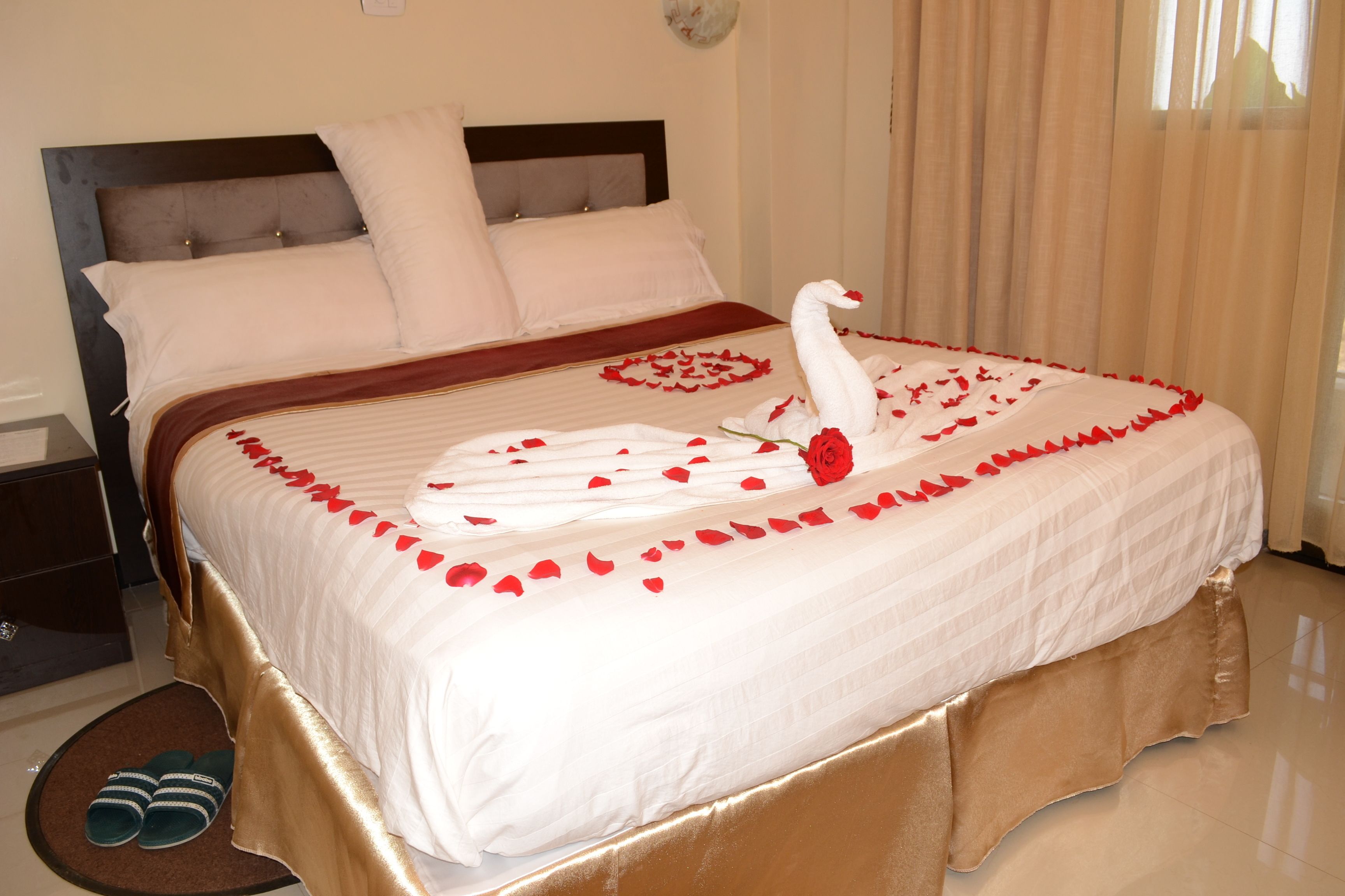 A neatly made bed with a swan and rose decorations, a pair of slippers, and a wooden nightstand.