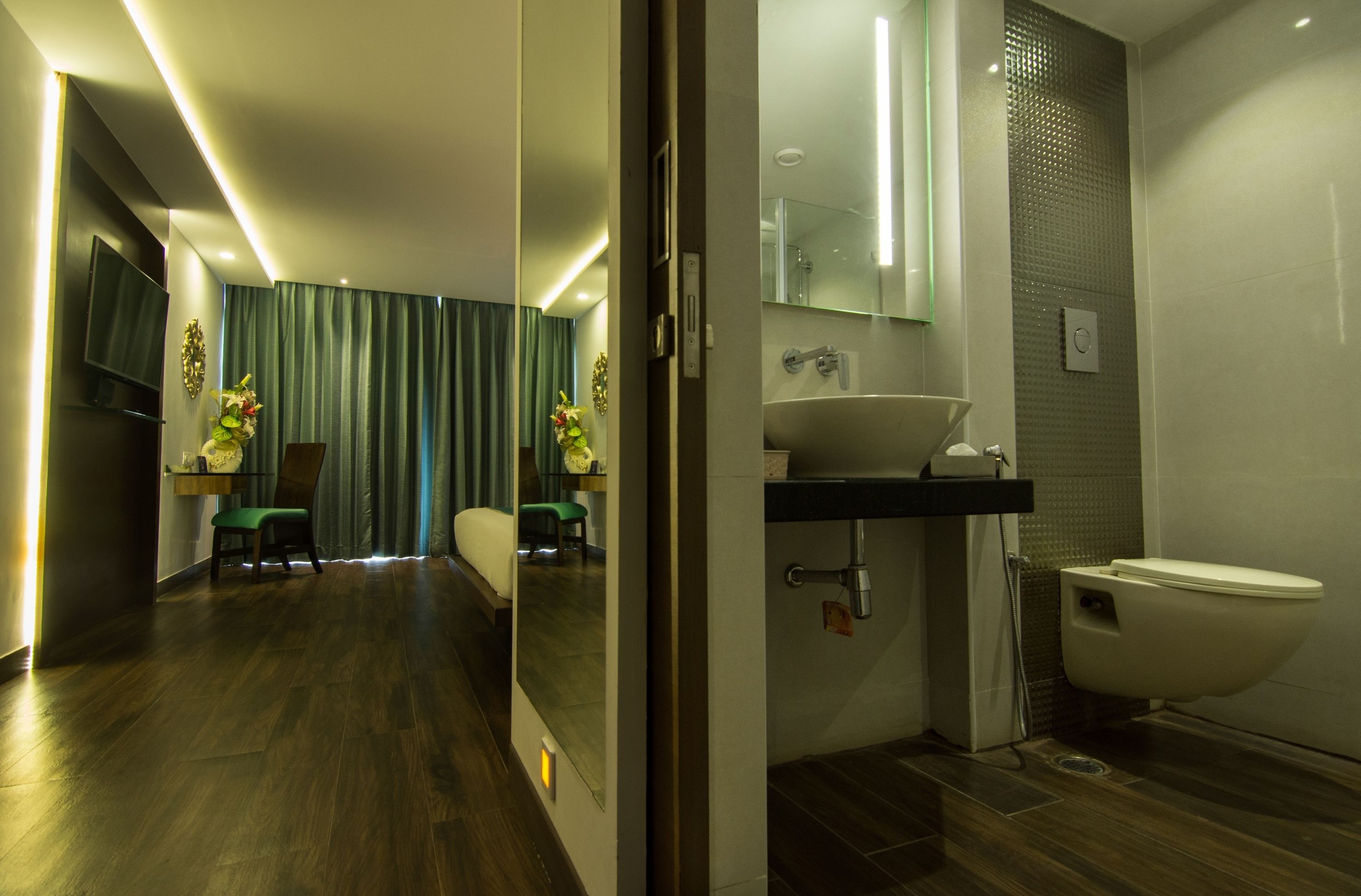 Un baño moderno con un espejo grande, un lavamanos y un inodoro. Cuenta con una zona de ducha con puerta de cristal y una pequeña área de descanso con una silla y una mesa.