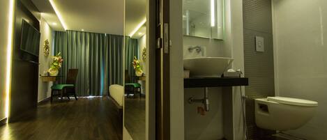 Un baño moderno con un espejo grande, un lavamanos y un inodoro. Cuenta con una zona de ducha con puerta de cristal y una pequeña área de descanso con una silla y una mesa.