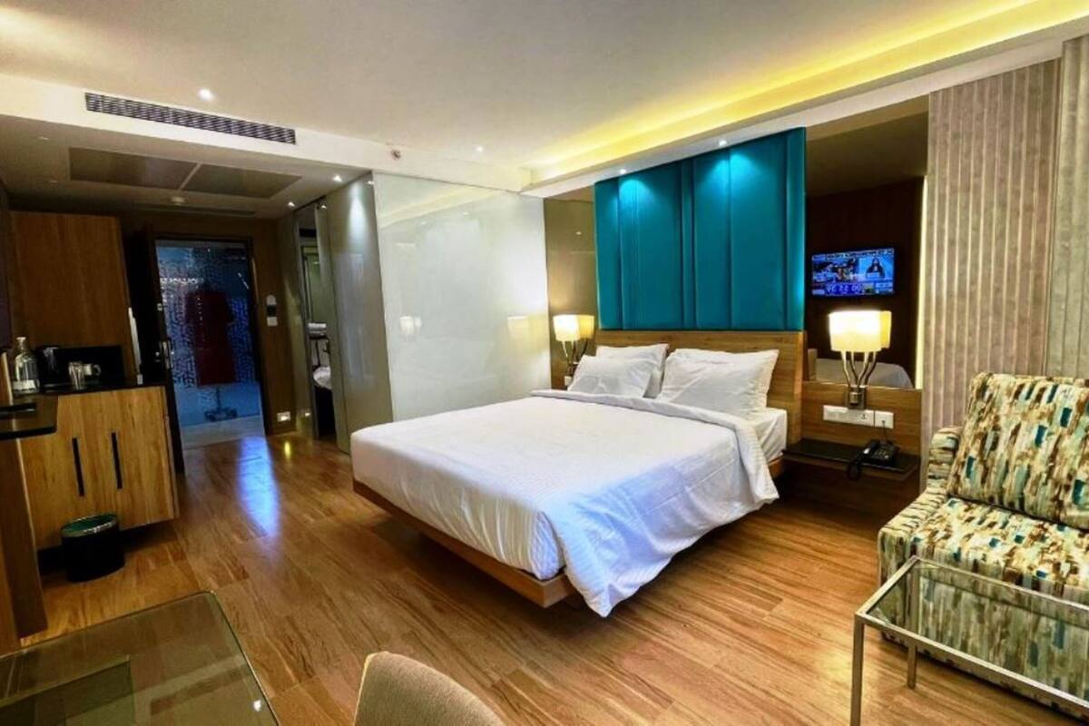 A modern hotel room with a large bed, a seating area with a patterned armchair, a TV, and a minibar.