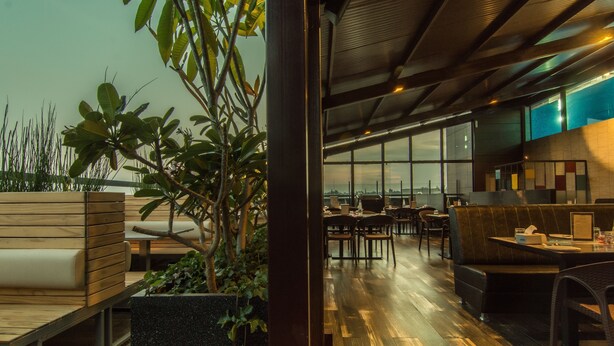 A modern indoor space with wooden seating, a large window offering a view, and a plant in the foreground. - Roopa Elite (Mysore)