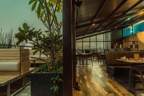 A modern indoor space with wooden seating, a large window offering a view, and a plant in the foreground. - Roopa Elite (Mysore)