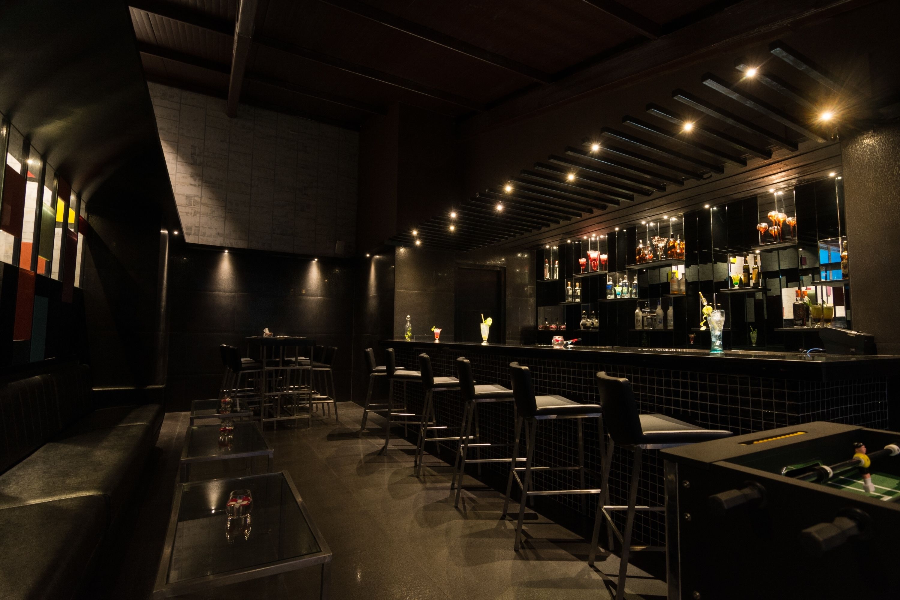 Un bar tenuemente iluminado con varios taburetes y mesas, una barra con diferentes botellas y copas, y un diseño moderno en el techo.