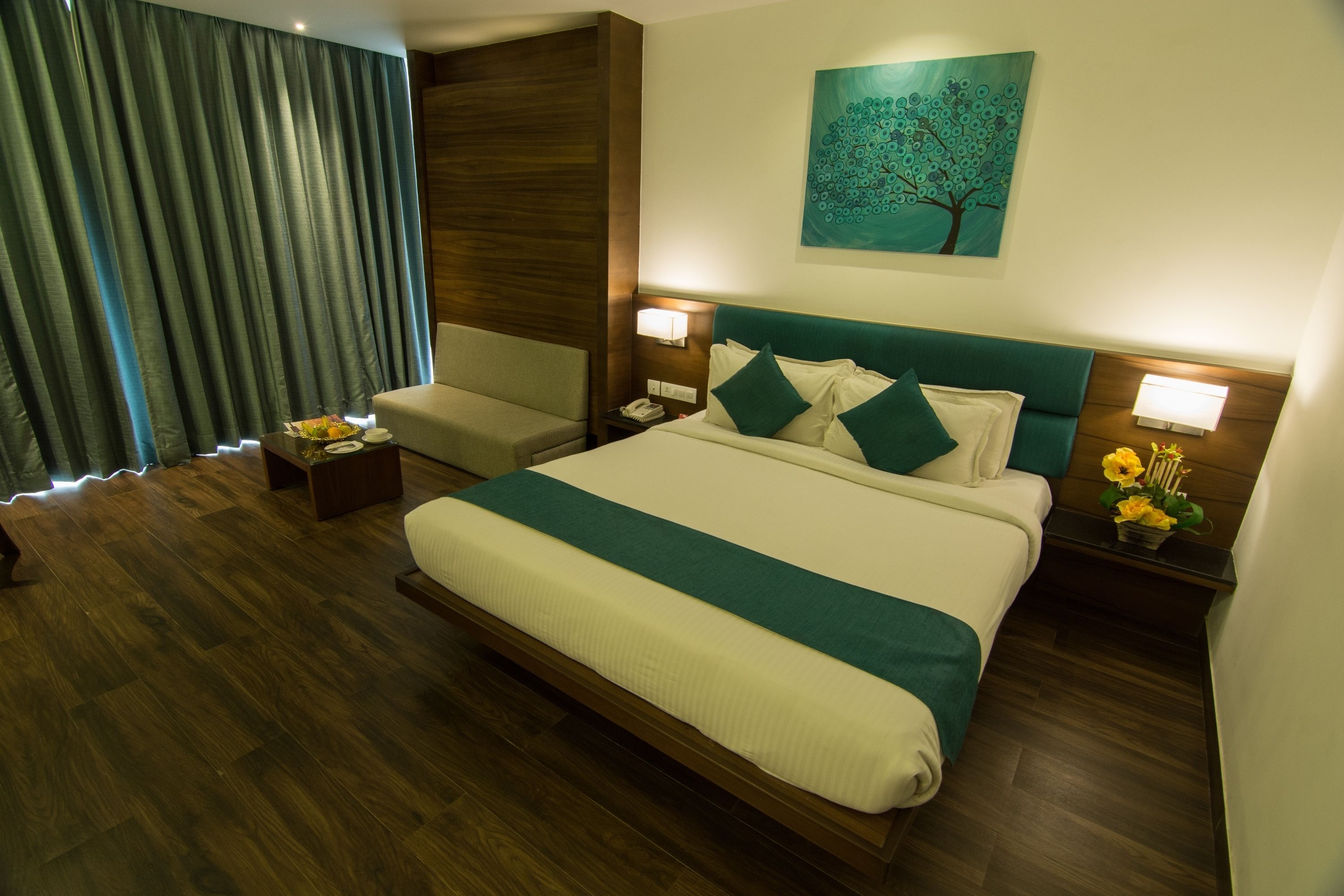 A hotel room with a large bed, a sitting area, wooden flooring, a painting on the wall, and a nightstand with a lamp.
