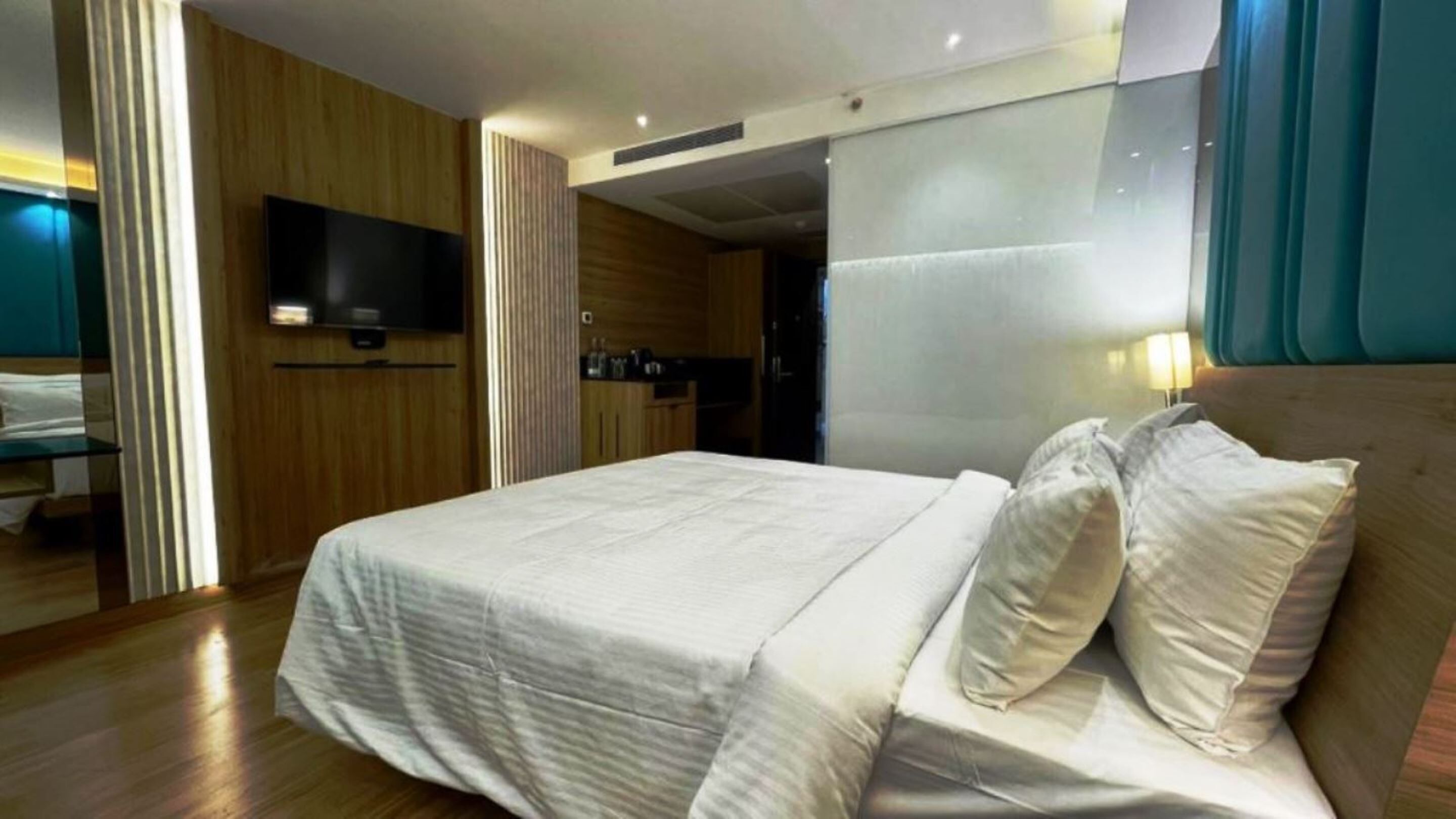 A modern hotel room with a large bed, a flat-screen TV mounted on the wall, a wooden panel wall, and a view of the bathroom.