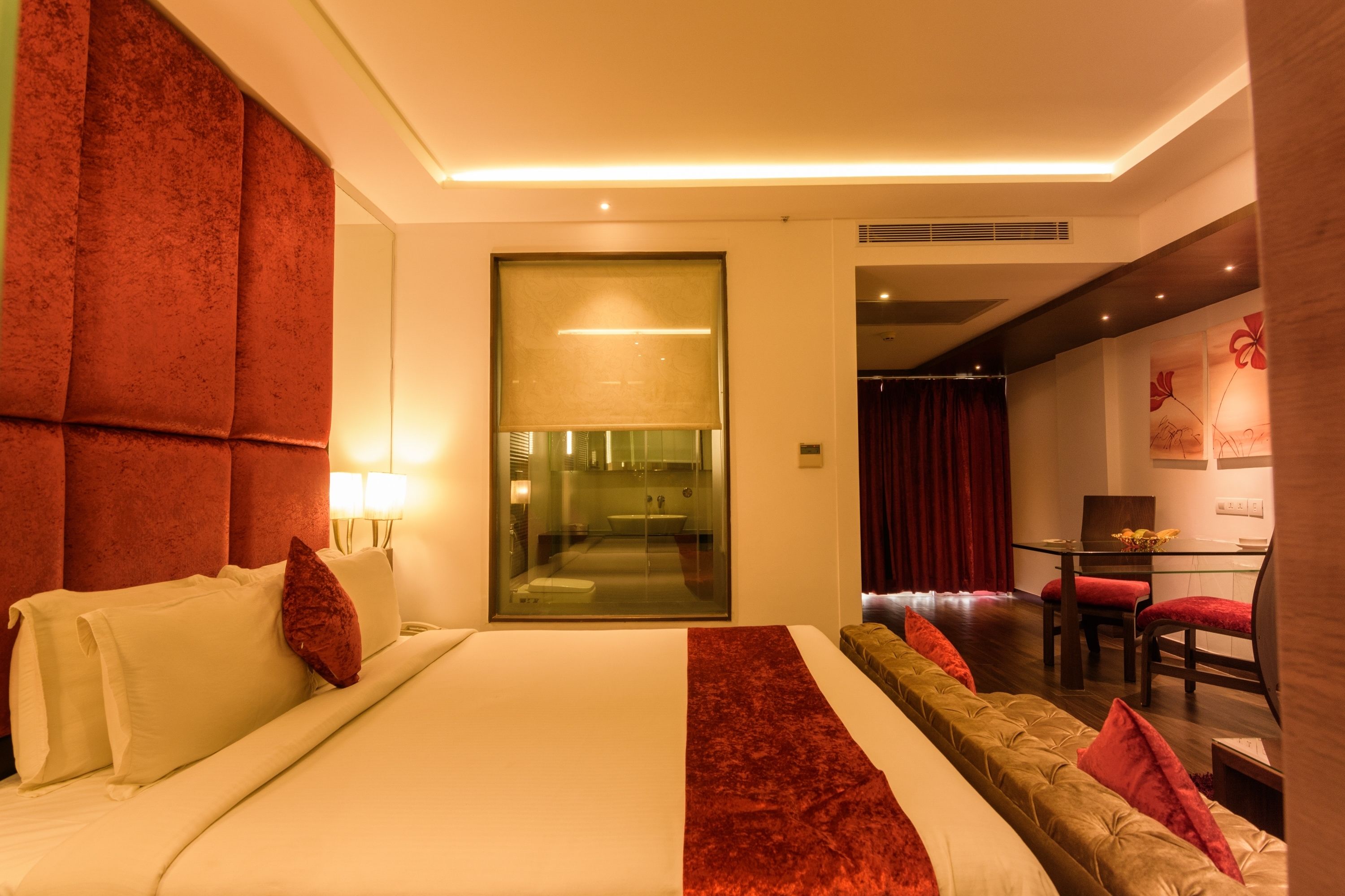 Una habitación de hotel moderna con una cama grande, un separador de vidrio, un área de comedor con mesa y sillas, y una cortina roja.