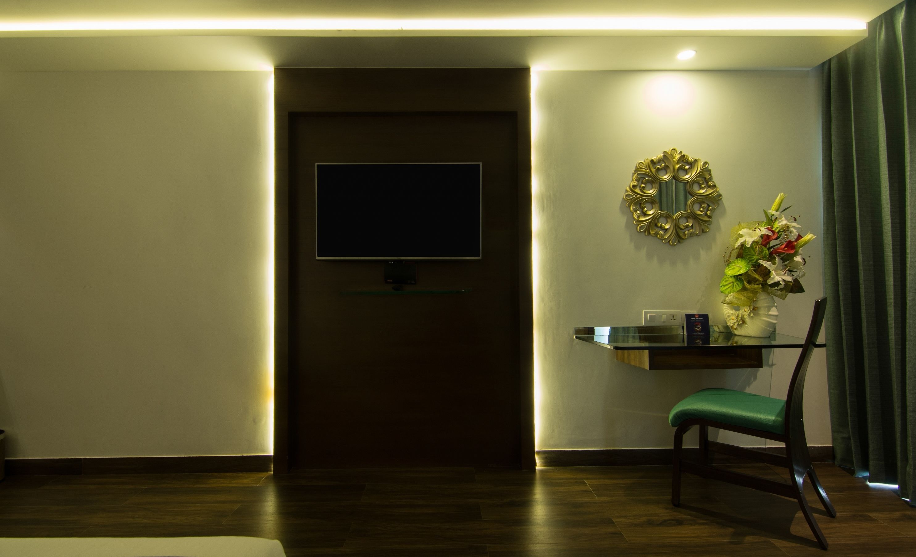 Habitación de hotel con televisor de pantalla plana montado en la pared, una mesita con un jarrón de flores y una silla tapizada en verde.