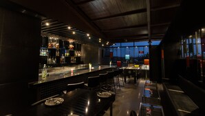 A modern bar area with a high ceiling, bar stools, and a view of the cityscape at dusk. - Roopa Elite (Mysore)