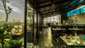 A restaurant with a view of the sky and plants, featuring wooden tables and chairs, and a modern interior design. - Roopa Elite (Mysore)