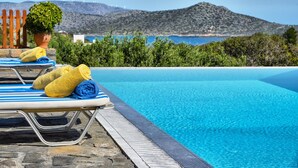 A poolside area with lounge chairs and towels, a potted plant, and a view of the sea and hills. - Glan Y Mor (Agios Nikolaos)