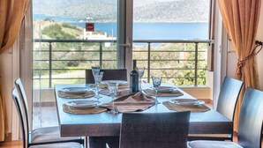 A dining table set with plates, glasses, and a bottle, overlooking a scenic view of a body of water and hills. - Glan Y Mor (Agios Nikolaos)