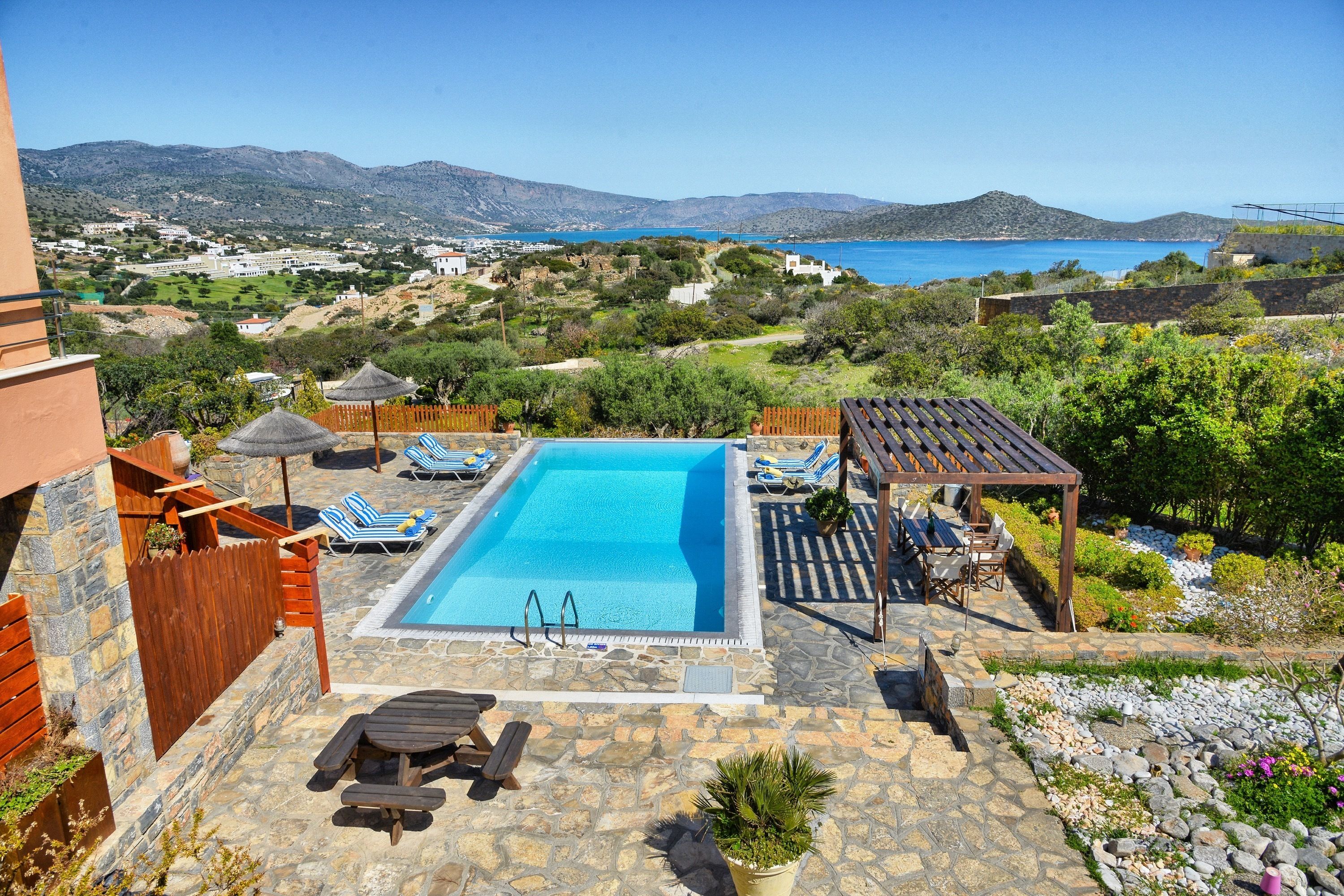 A swimming pool with lounge chairs and a pergola, surrounded by stone tiles and greenery, overlooking a coastal landscape.