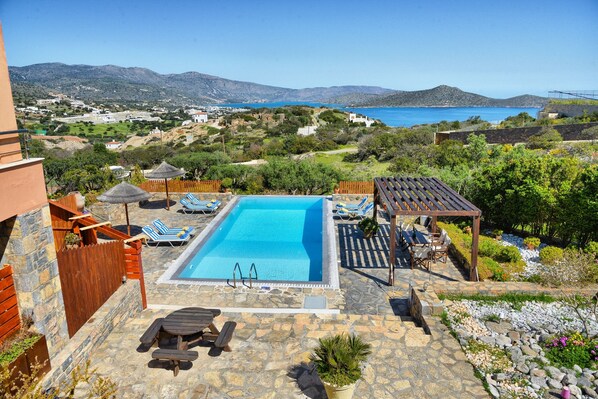 A swimming pool with lounge chairs and a pergola, surrounded by stone tiles and greenery, overlooking a coastal landscape.