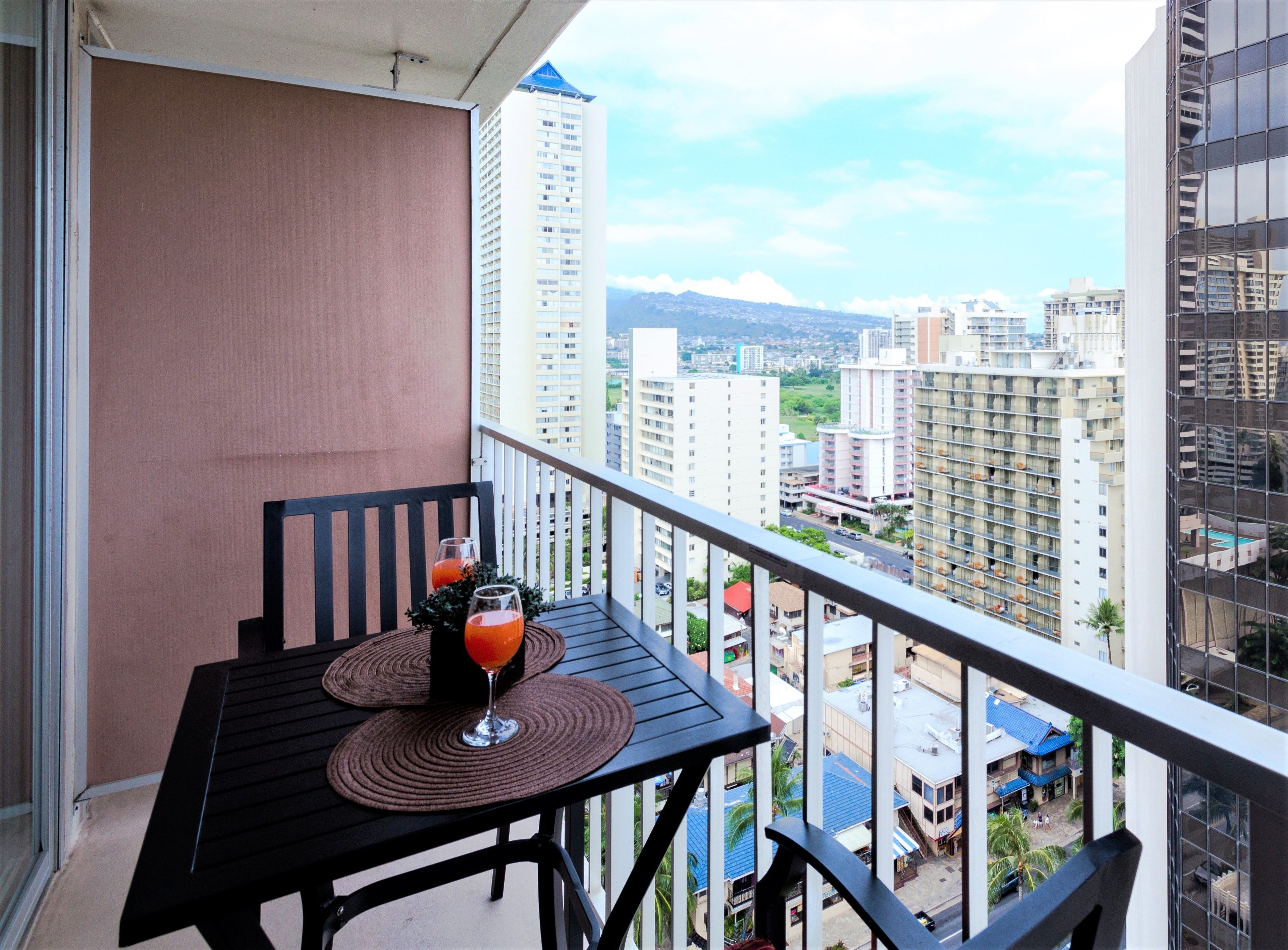 A balcony with a table and two glasses of orange juice, overlooking a cityscape with tall buildings.