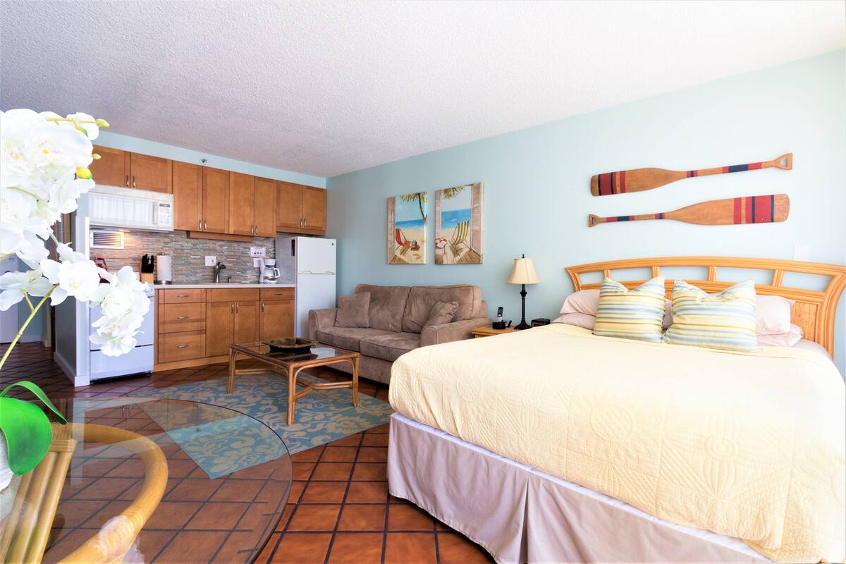 A compact room with a kitchenette, a bed with a beige quilt, a wooden headboard, and two wall-mounted paddleboards.