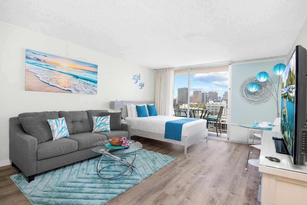 A modern hotel room with a city view, a sofa, a bed, and a dining area.