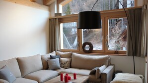 A cozy living room with a beige sectional sofa, a wooden coffee table with red candles, and a large window with a view of a snowy landscape. - Chalet Binna (Zermatt)