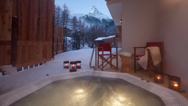A hot tub with a snowy mountain view, wooden deck, and two red chairs. - Chalet Binna (Zermatt)