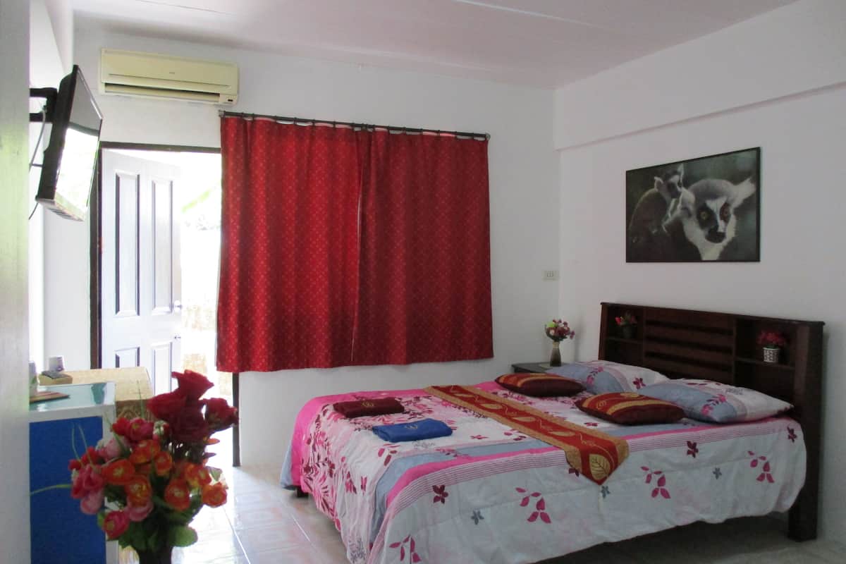 Ein Schlafzimmer mit einem Bett, einem Fenster mit roten Vorhängen, einem an der Wand montierten Fernseher, einem Gemälde und einer Vase mit Blumen.