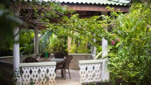 Un espace extérieur aménagé, abrité et doté d’une table et de chaises, entouré d’une végétation luxuriante et d’une clôture blanche en treillis.