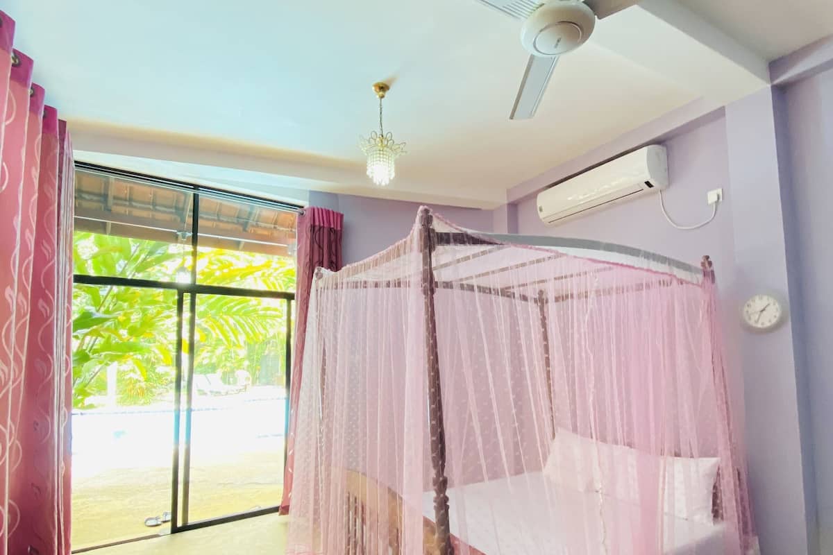 Quarto com cama de dossel, ventilador de teto, aparelho de ar condicionado e vista para o verde pela janela.