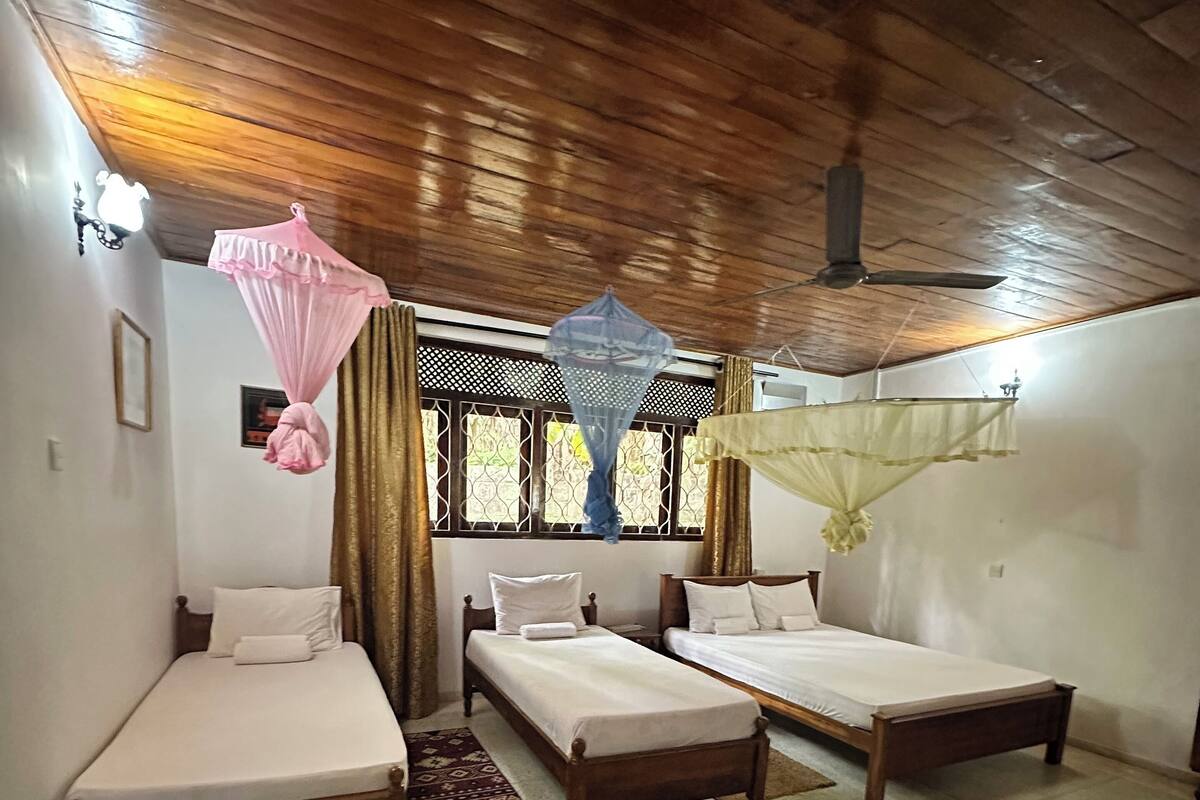 Quarto com duas camas de madeira, um ventilador de teto e uma janela com cortinas.
