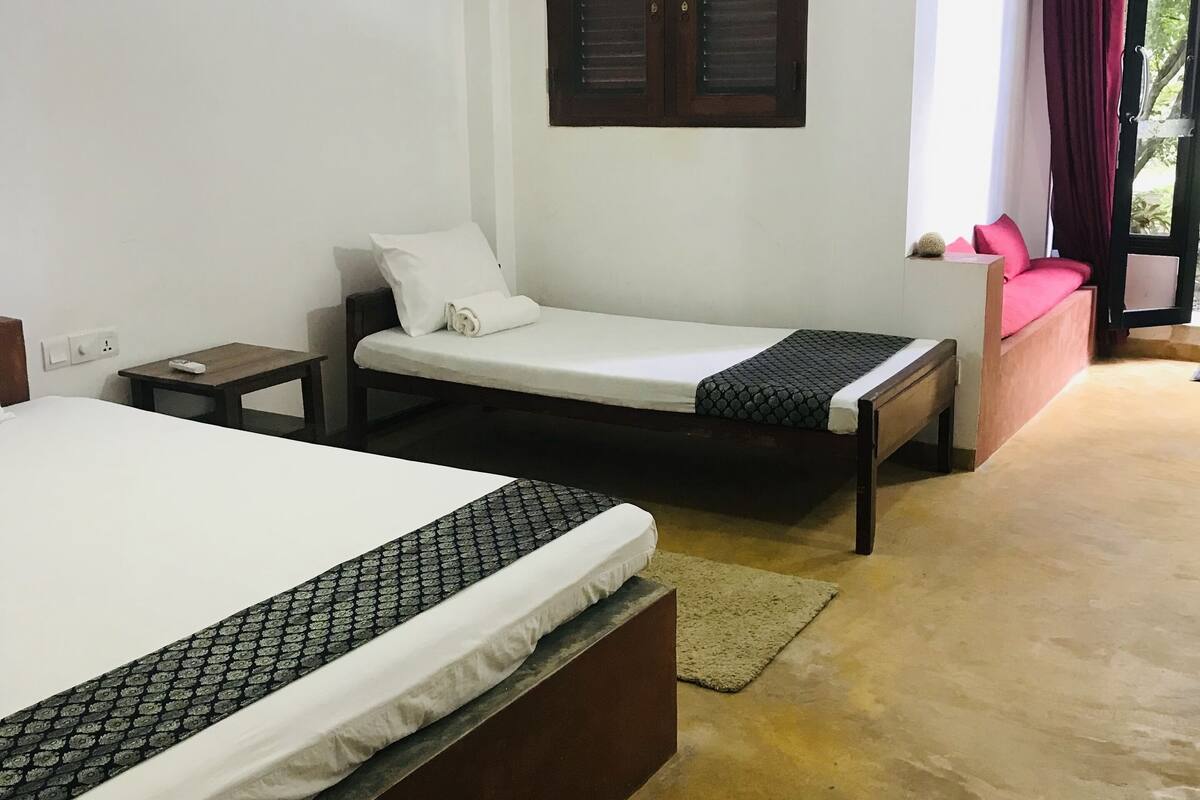 Quarto de hotel com uma cama grande, uma cama menor, um banco, uma mesa de cabeceira, uma janela decorativa e vista para o verde lá fora.