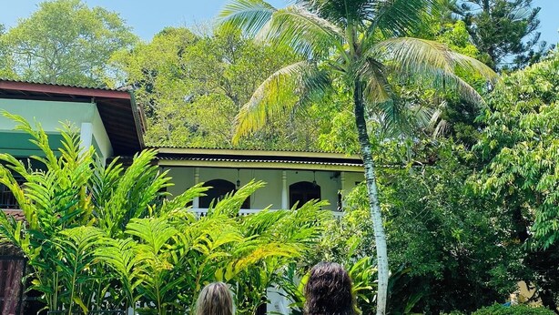 Deux personnes sont assises au bord d’une piscine, avec une maison et une végétation tropicale en arrière-plan.