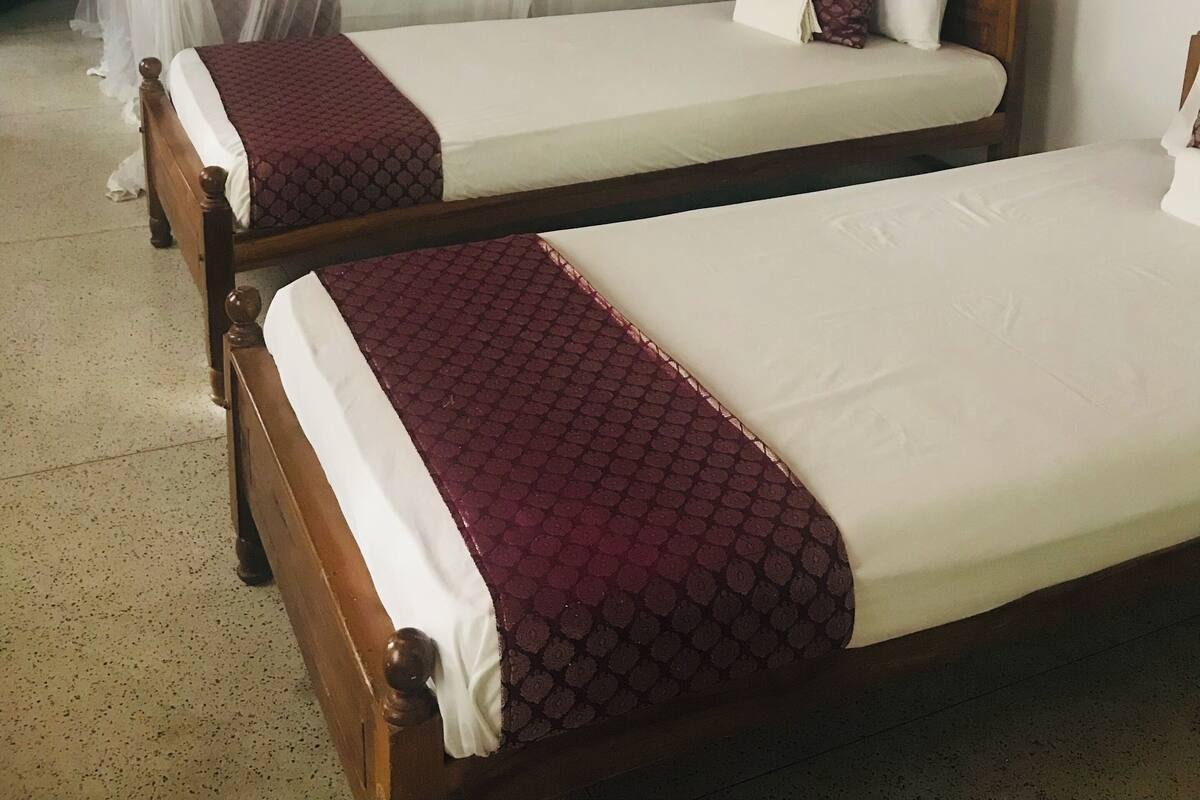 Duas camas de madeira com roupa de cama branca e bordô, teto de madeira e parede branca com uma pequena estátua.