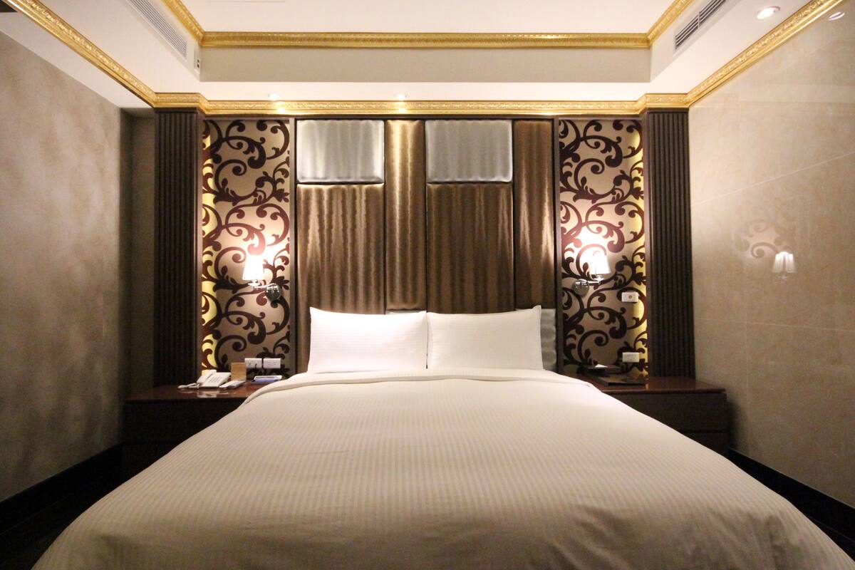 A hotel room with a large bed, two bedside tables, a nightstand, a decorative headboard, and two wall-mounted lamps.