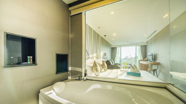 A modern bathroom with a large bathtub, a small shelf with toiletries, and a large mirror reflecting the room. - Ratana Patong Beach Hotel by Shanaya (Patong)