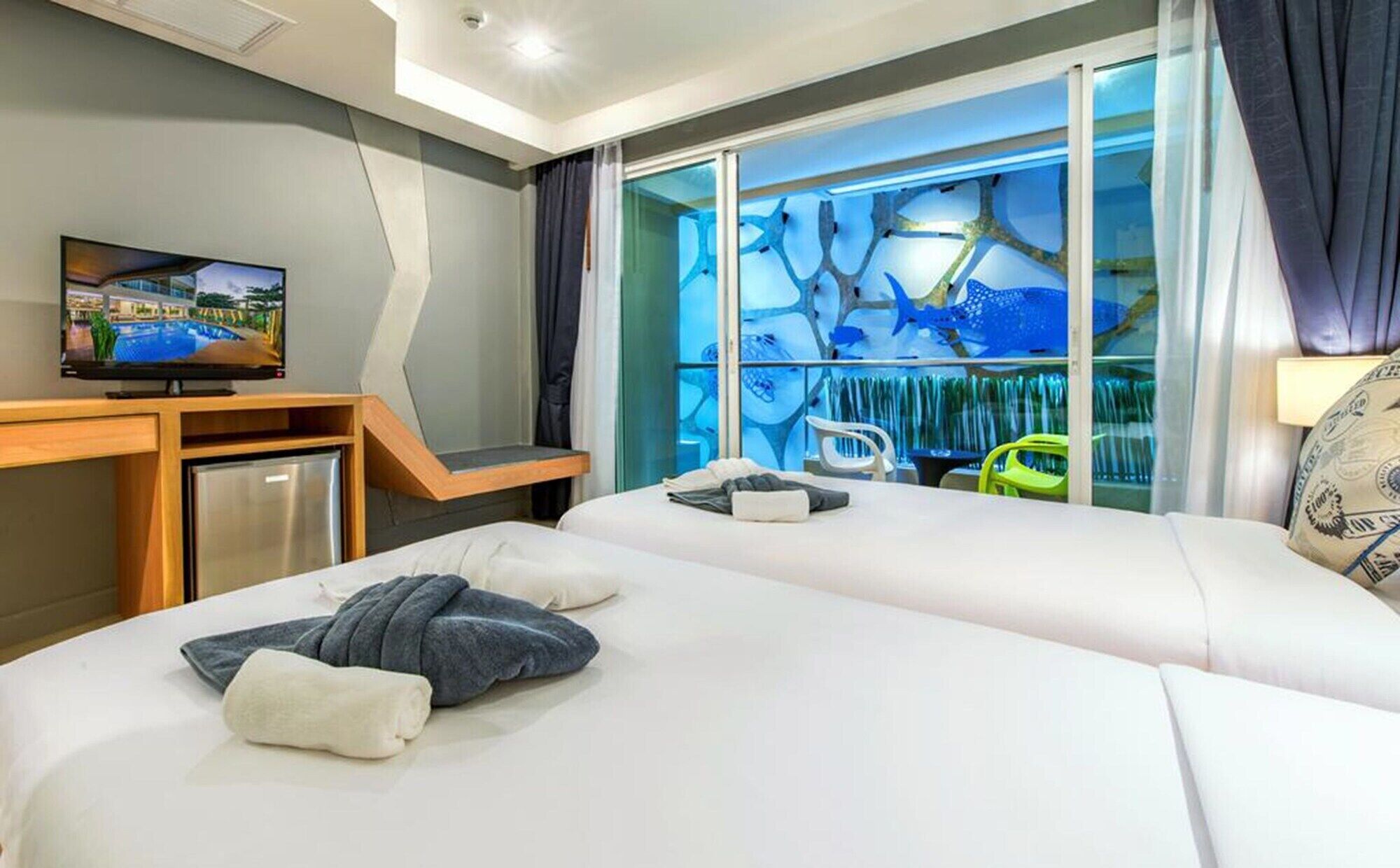 A hotel room with two beds, a TV, a desk, and a view of a blue aquatic scene through the glass wall.