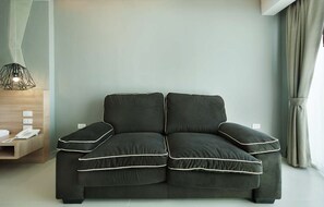 A modern living room with a dark grey sofa, a wooden side table with a telephone, and a geometric pendant light. - Ratana Patong Beach Hotel by Shanaya (Patong)