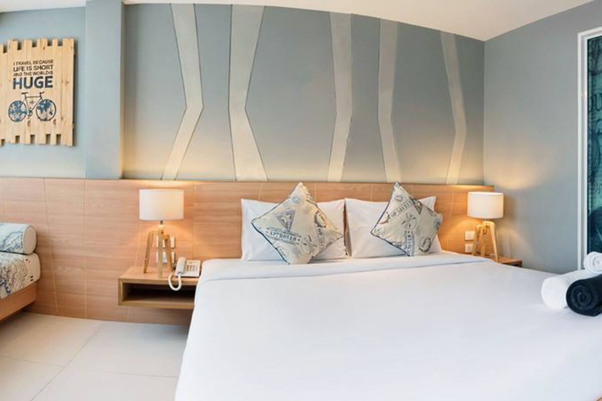 Habitación de hotel moderna con una cama grande, un sofá más pequeño y una mesita de noche con lámpara. Se aprecia una pared con diseño de mapa y una puerta decorada con motivos de viaje.
